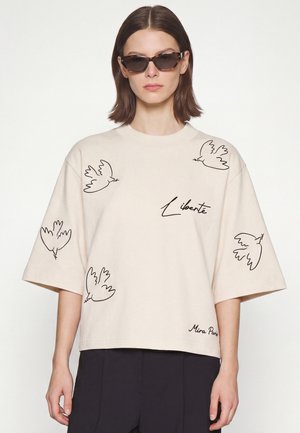 Femme portant une chemise beige oversize avec des croquis d'oiseaux noirs et les textes "Liberté" et "Mira Paris,", associée à un pantalon foncé et des lunettes de soleil.