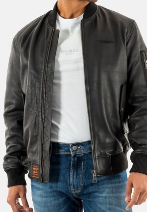 Homme portant une veste bomber en cuir noire sur un t-shirt blanc et un jean bleu, avec la veste dézippée montrant le logo du t-shirt.