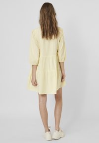Gul, knälång klänning med flerlagersdesign, puffärmar och avslappnad passform. Tillverkad av mjukt tyg och har en enkel bakknäppning.