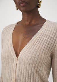 Cardigan côtelé beige clair avec col en V, présentant une texture douce et deux boutons crème à l'avant. Détails subtils le long des bords.