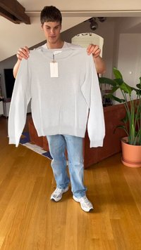 Giovane uomo in piedi all'interno che tiene un maglione lavorato a maglia grigio chiaro con un'etichetta davanti a un divano marrone e a una pianta verde in vaso.