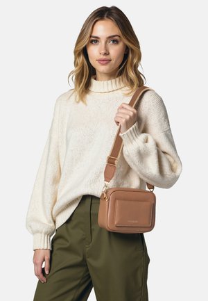 Tasche aus braunem Leder im Querformat mit quadratischer Form, Reißverschluss und Fronttasche; Modell trägt einen cremefarbenen Pullover und olivgrüne Hose.