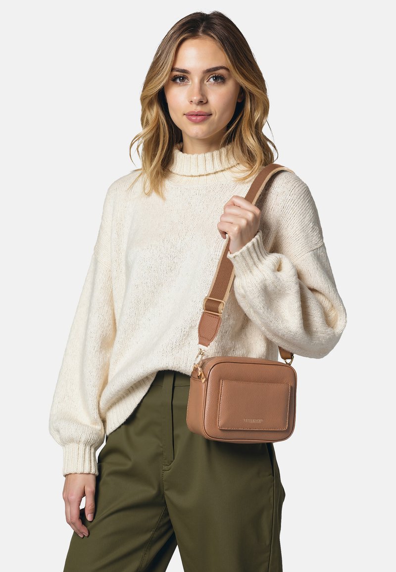 Borsa a tracolla in pelle tan con forma quadrata, chiusura con zip e tasca anteriore; modello che indossa un maglione crema e pantaloni verde oliva.