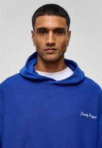 Blauwe fleece hoodie van een gestructureerde stof, met een grote capuchon en een klein wit logo op de borst. Gelaagd over een wit shirt.