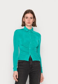 Camisa azul-turquesa de manga longa, abotoada, com gola e detalhe franzido na parte da frente, apresentando uma textura suave. Estilizada com jeans pretos.