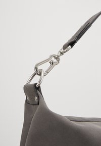 Gros plan sur une lanière de sac grise attachée au sac avec un mousqueton argenté et un anneau métallique sur un fond uni.