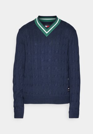 Man draagt een donkerblauwe kabelgebreide V-hals trui en gebroken witte cargobroek met witte sneakers, staand op een effen achtergrond.