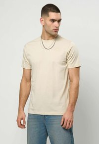 Beige bomull T-skjorte med rund hals, korte ermer og en liten logo på brystet; kombinert med lyseblå jeans. To sølvkjeder er synlige.