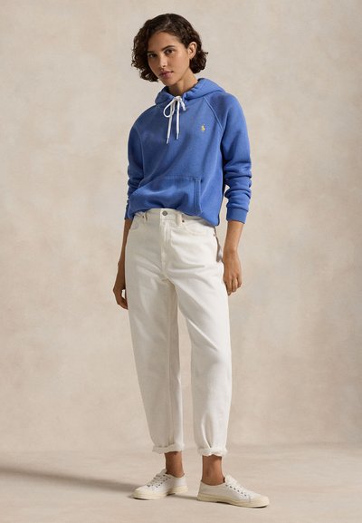 Moteris vilki mėlynį gaubtu džemperį su baltais raišteliais, baltais užriestais kelnėmis ir baltais sportbačiais, stovinti priešais paprastą foną.