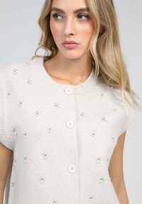 Cardigan en tricot beige à manches courtes, avec une broderie florale décorative, deux boutons blancs à l'avant et une texture douce.