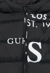 Doudoune noire avec fermeture éclair, ornée d'un grand texte blanc et d'un logo triangulaire. Le matériau semble lisse avec une texture matelassée.