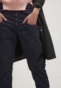 Granatowe spodnie o gładkiej fakturze, z pięcioma guzikami na pasku i kieszeniami z przodu, noszone z jasnoróżowym swetrem.