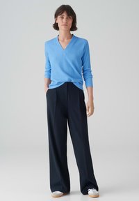 Hellblaue, strukturierte V-Ausschnitt-Strickjacke kombiniert mit schwarzen, weit geschnittenen Hosen und weißen Sneakers. Einfaches, modernes Outfit vor einem neutralen Hintergrund.