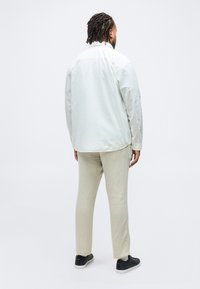 Άνδρας με πλεξουδάκια στέκεται με την πλάτη προς το μέρος μας, φορώντας λευκό-κρεμ πουκάμισο, μπεζ παντελόνι, ανοιχτόχρωμες κάλτσες και μαύρα αθλητικά παπούτσια με λευκές σόλες.