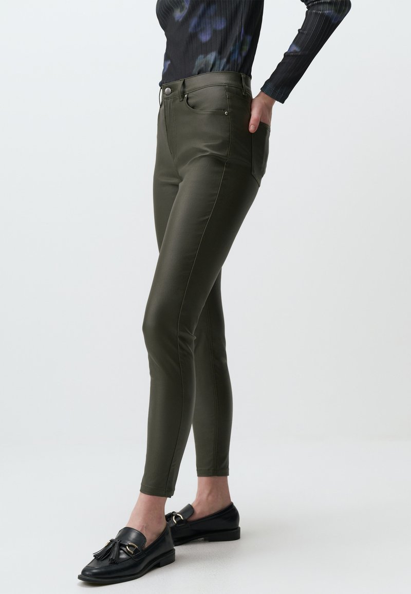 Olijfgroene, nauw aangesloten broek met een glanzende afwerking, middelhoge taille, voorzakken en subtiele stikdetails. Gecombineerd met zwarte instapschoenen.