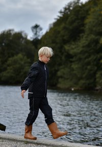 Black quilted jacket with a zip, black pants, and tan waterproof boots on a child walking beside a body of water with greenery in the background.