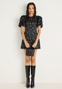 Crna haljina s puf rukavima, cvjetnim uzorkom, suženim strukom i detaljem u obliku ključanice. Uparena s crnim čizmama do koljena i torbom s potkovicama.