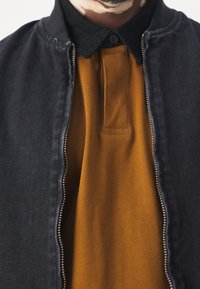 Czarna kurtka bomber z denimu z zamkiem błyskawicznym, noszona na musztardowej koszulce polo. Kurtka ma zaokrąglony kołnierz i fakturowaną tkaninę.