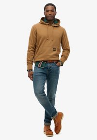 Felpa con cappuccio marrone e tasca sul davanti, indossata sopra una camicia a quadri verde, abbinata a jeans blu e stivali marroni con lacci.