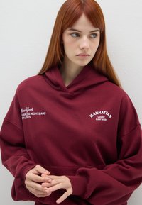 Giovane donna con lunghi capelli rossi che indossa un hoodie bordeaux con la scritta "Manhattan Upper East Side" e "New York Notti Insonni e Luci della Città".