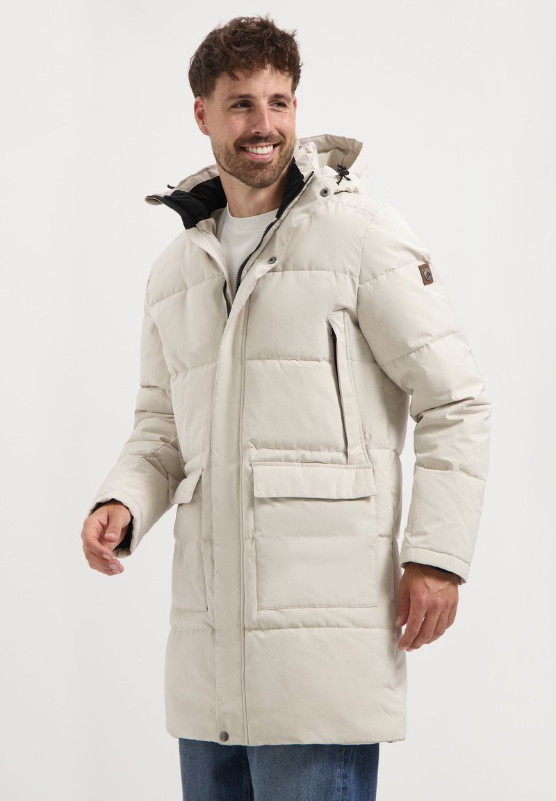 Glimlachende man in een lichtbeige gewatteerde winterjas met capuchon en grote voorzakken, staand tegen een eenvoudige achtergrond.