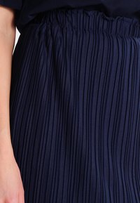 Zbliżenie dłoni kobiety spoczywającej na granatowej plisowanej spódnicy, ukazujące teksturowany materiał i elastyczny pas.