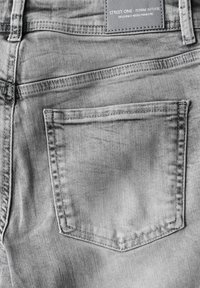 Tejido de mezclilla gris con un bolsillo trasero que presenta detalles de costura visibles. La etiqueta dice "Street One - Denim Atelier." Textura suave.
