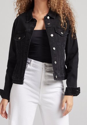 Kvinde med krøllet hår bærer en sort denimjakke over en sort top og højtaljede hvide jeans, stående mod en neutral baggrund.