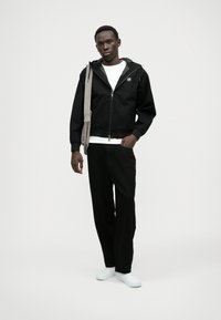 Čierna bunda na zips s kapucňou, oblečená na bielom tričku. Skombinovaná s čiernymi nohavicami a bielymi teniskami, doplnená svetlošedým popruhom na taške.