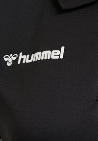 Logo del marchio Hummel bianco e ape stilizzata stampati su tessuto nero texturizzato, probabilmente abbigliamento sportivo.