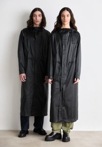 Sorte vandtætte trenchcoats lavet af glat materiale, med hætte, forlommer og lige skæring, båret over løstsiddende mørke bukser.