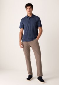 Tengerészkék rövid ujjú ing finom mintával, világos bézs nadrággal és sötét sportcipővel párosítva, semleges háttér előtt állva.