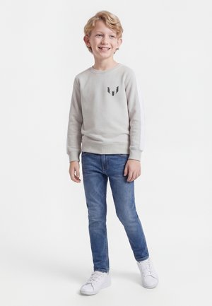 Χαμογελαστό αγόρι με ξανθά μαλλιά που φοράει ανοιχτό γκρι φούτερ, μπλε τζιν και λευκά αθλητικά παπούτσια, στέκεται μπροστά σε λευκό φόντο.