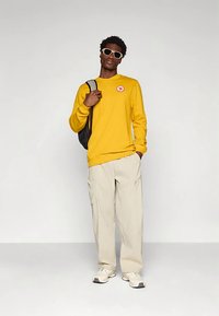 Żółta koszula z długim rękawem z naszywką z logo, zestawiona z beżowymi spodniami o szerokich nogawkach i białymi trampkami; do stroju dodano czarny plecak oraz okulary przeciwsłoneczne.