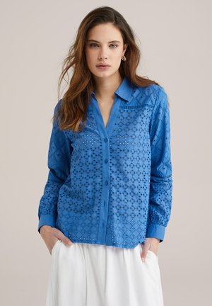 Camicia blu con bottoni, caratterizzata da intricatissimi motivi a intaglio, maniche lunghe e colletto. Il tessuto sembra leggero e traspirante.