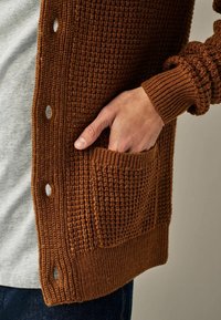 Brązowy dziergany kardigan z teksturowanym wzorem, z boczną kieszenią, ściągaczami na rękawach i zapinany na guziki z przodu.