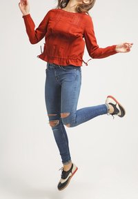 Haut rouge à manches longues avec des détails texturés et un ourlet à cordon de serrage, associé à un jean skinny bleu déchiré et des chaussures noir et blanc.