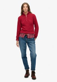 Czerwony sweter z półzamkiem, noszony na koszuli w kratę. Do tego niebieskie dżinsy i brązowe sznurowane botki.