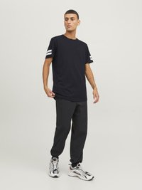 Camiseta negra de algodón con rayas horizontales blancas en las mangas, combinada con pantalones negros ajustados y zapatillas deportivas multicolores.