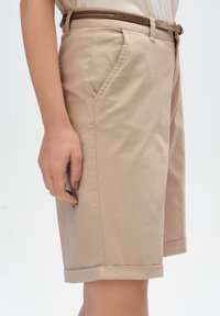Beżowe bawełniane szorty z bocznymi kieszeniami, z podwiniętym dołem i brązowym paskiem. Charakteryzują się gładką fakturą i dopasowanym krojem.