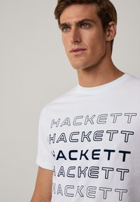 Camiseta de algodón blanca con el texto "HACKETT" repetido en diferentes grosores de fuente. Cuello redondo y mangas cortas. Textura suave.