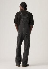 Άνδρας με πλεξούδες, φοράει μαύρο υπερμεγέθες μπλουζάκι και άνετο σκούρο γκρι τζιν φορμάκι, στέκεται με την πλάτη προς το φακό σε απλό φόντο.