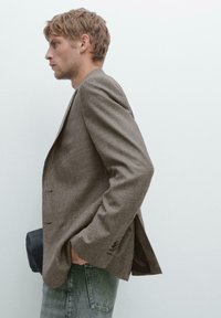 Brown houndstooth blazer with two buttons, side vents, and textured fabric, worn over gray denim jeans. Simple, modern design.