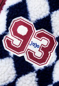 Tekstūrēta balta auduma ar tumši zilu un pūkainu rakstu; piedāvā bordo izšūtu uzlīmi, kas parāda "93" un "Jojo" zilā krāsā.