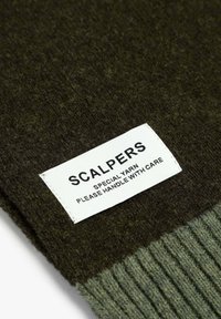 Tejido de punto verde con una textura acanalada, que cuenta con una etiqueta blanca que dice "SCALPERS SPECIAL YARN, MANEJAR CON CUIDADO".