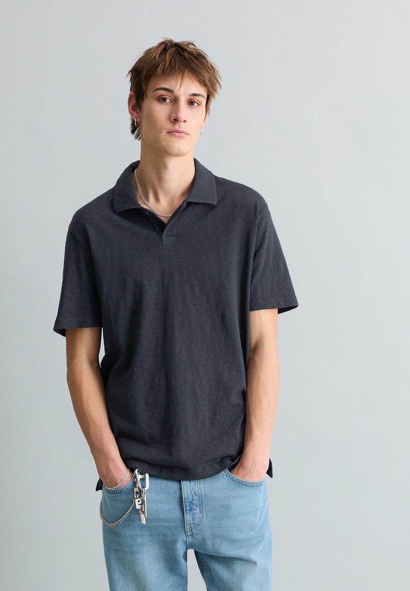 Jeune personne aux cheveux bruns courts portant un polo gris foncé et un jean bleu clair avec des clés accrochées à la boucle de ceinture, debout devant un fond gris.