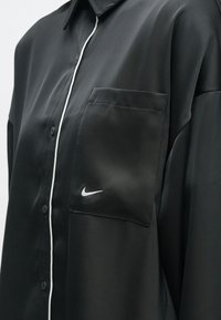 Μαύρο πουκάμισο με κουμπιά, με λευκή ραφή κατά μήκος της ραφής των κουμπιών και μια τσέπη στο στήθος που διαθέτει ένα μικρό λευκό λογότυπο νίκης της Nike.