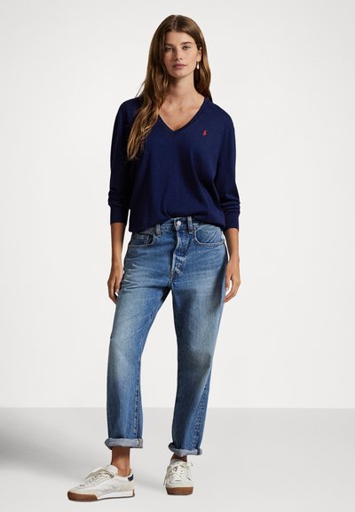 Camisola azul-marinho com decote em V, ajuste descontraído, combinada com jeans de perna reta em lavagem clara, bainhas dobradas, e ténis brancos com solas castanhas.