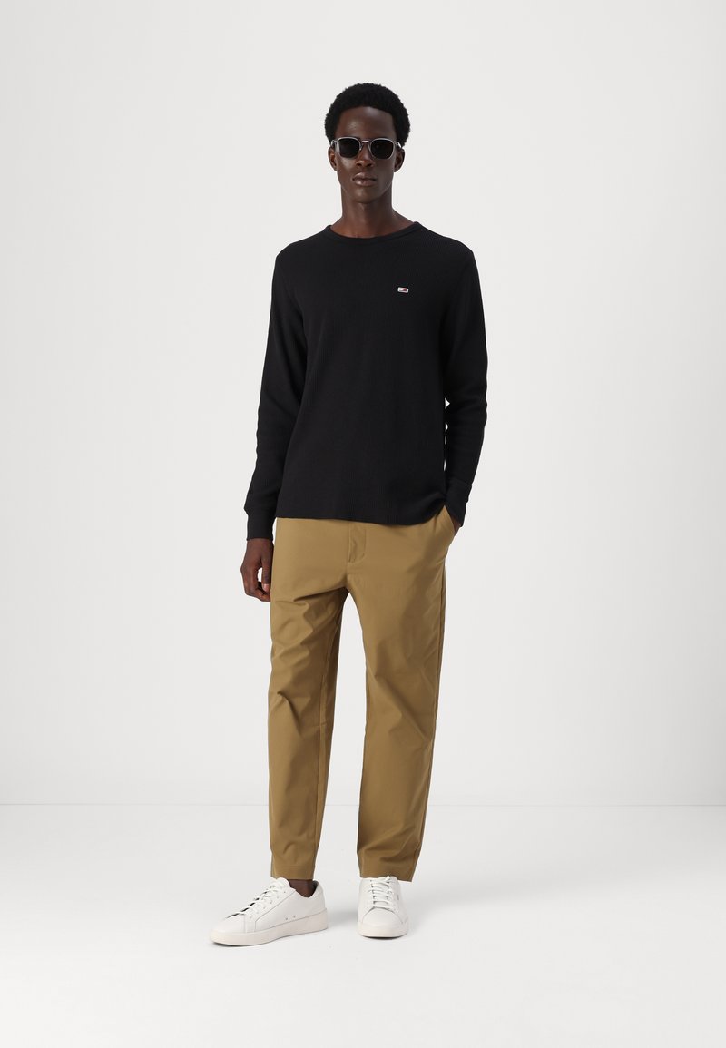 Czarna koszula z długim rękawem, beżowe spodnie, białe trampki. Koszula ma małe logo. Model nosi okulary przeciwsłoneczne i stoi na jednolitym tle.