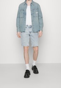 Camicia di denim azzurro chiaro con due tasche sul petto, maglietta bianca, pantaloncini di denim azzurro chiaro, calze bianche e scarpe slip-on nere.